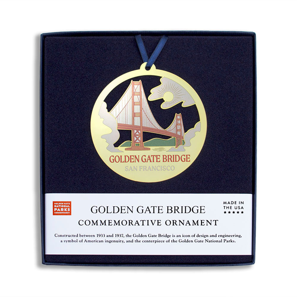 Round gold ornament with multi-color cutout design of San Francisco Golden Gate Bridge, packaged in square box with blue velvet insert